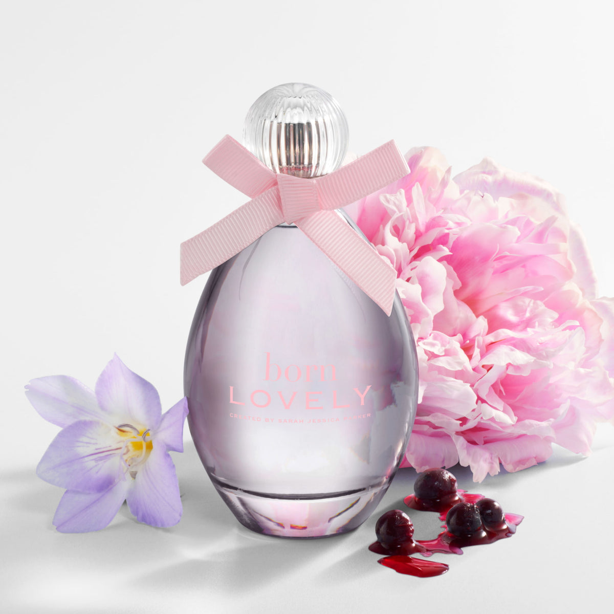 A perfume bottle labeled 'born LOVELY' with a pink ribbon bow, styled with a pink peony flower, a purple iris, and red cherries on a clean white background.