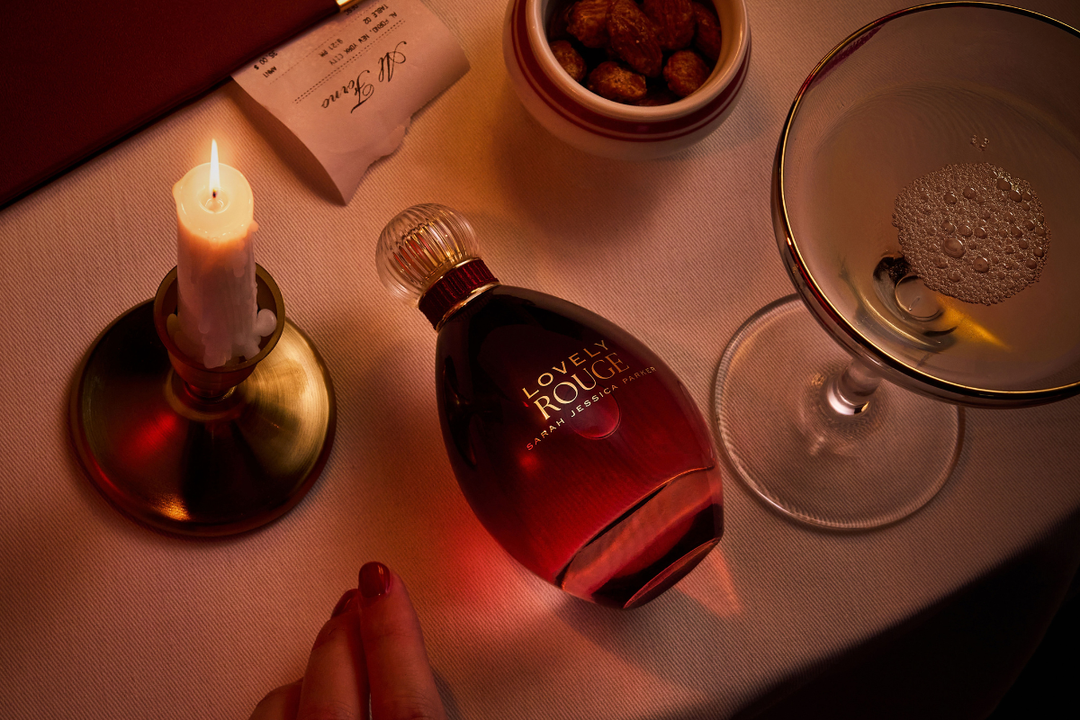 A bottle of Lovely Rouge Eau de Parfum lies on a table next to a lit candle in a brass holder, a cocktail glass, and a small bowl of nuts, with a hand resting near the bottom edge.