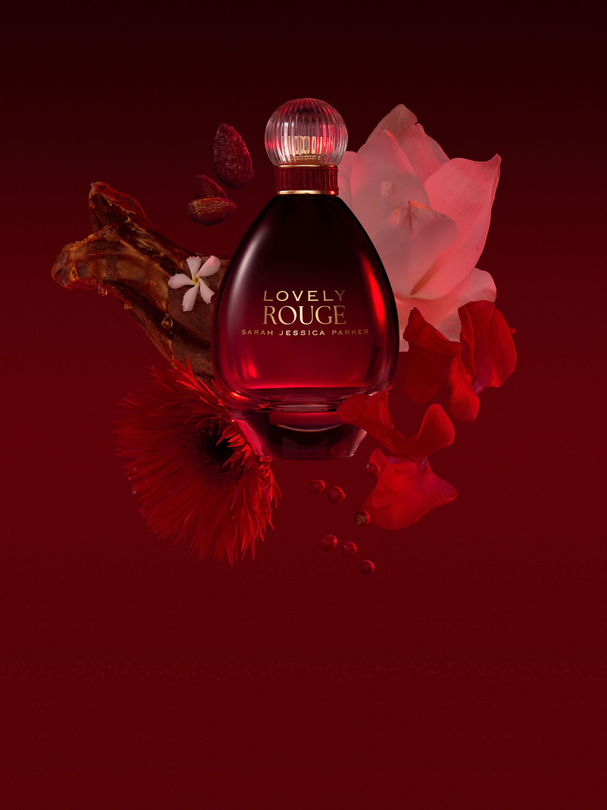A bottle of Sarah Jessica Parker's 'Lovely Rouge' perfume is centered against a dark red background, surrounded by red and pink flowers and a dried leaf.