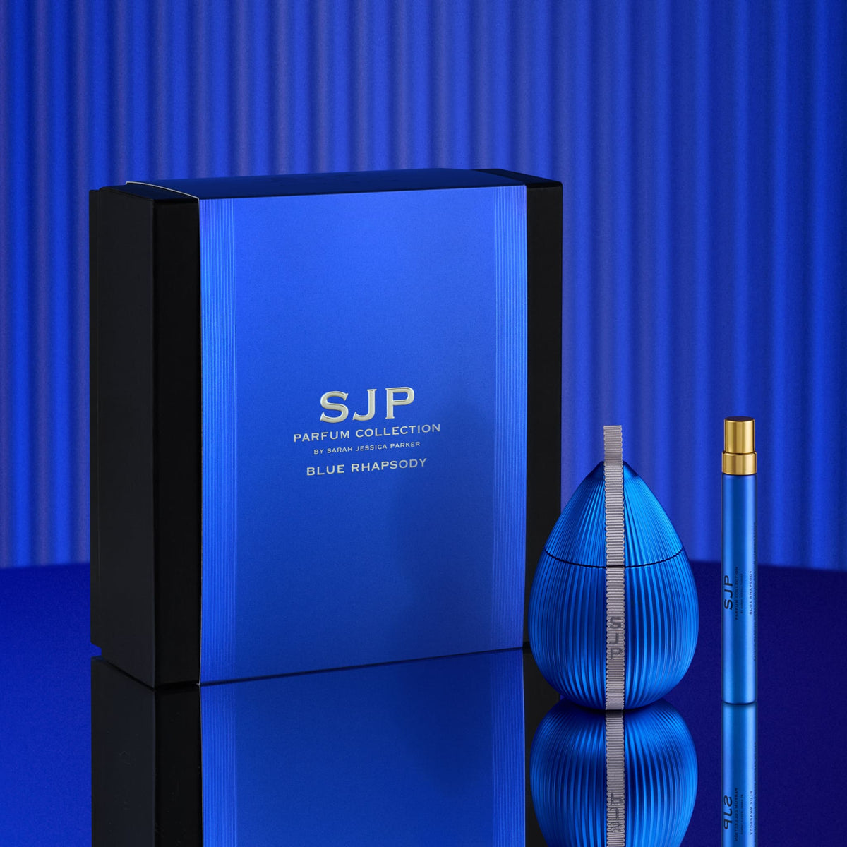 Blue Rhapsody perfume set by SJP Parfum Collection, featuring a large blue and black box, a ribbed blue bottle, and a matching blue travel spray vial with a gold cap, set against a blue striped background.