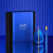 Blue Rhapsody perfume set by SJP Parfum Collection, featuring a large blue and black box, a ribbed blue bottle, and a matching blue travel spray vial with a gold cap, set against a blue striped background.