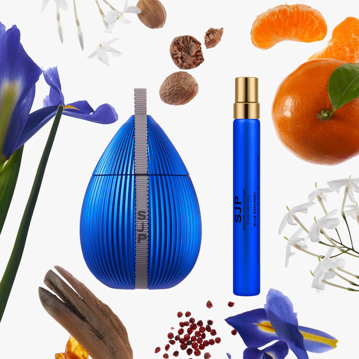 A blue teardrop-shaped perfume bottle with a vertical silver measurement scale design and a separate matching blue spray atomizer with a gold cap, arranged on a white background with floating botanical and citrus props including blue flowers, orange slices, nuts, and spices.