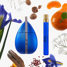 A blue teardrop-shaped perfume bottle with a vertical silver measurement scale design and a separate matching blue spray atomizer with a gold cap, arranged on a white background with floating botanical and citrus props including blue flowers, orange slices, nuts, and spices.