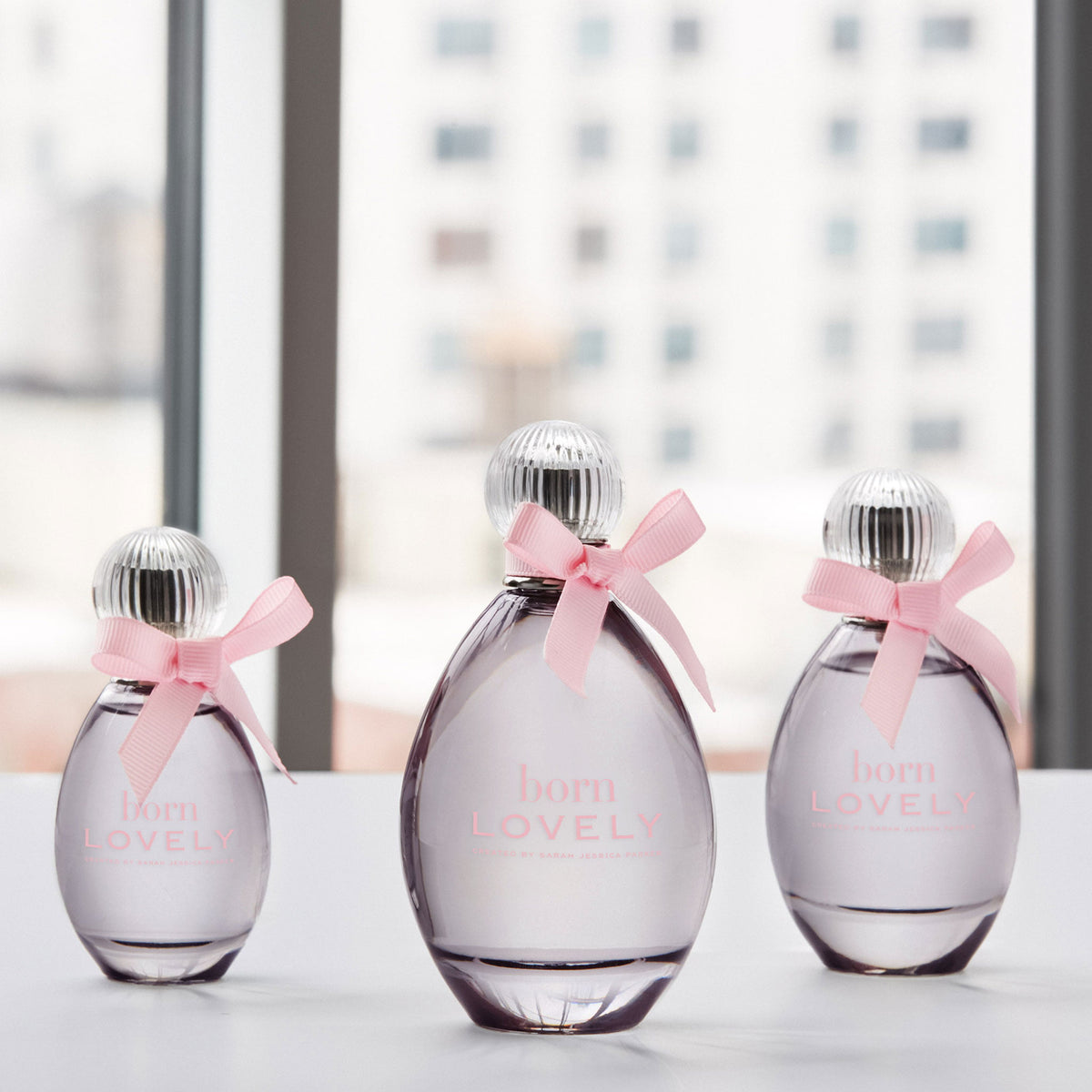 Three glass perfume bottles with silver caps and pink ribbons tied around the necks, labeled 'born LOVELY', arranged on a white surface in front of a softly blurred window background.