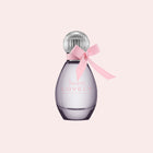 A bottle of Born Lovely Eau de Parfum by Chanel, featuring a translucent lavender-purple glass bottle, a silver ribbed cap, and a light pink satin ribbon tied in a bow around the neck, set against a solid pale pink background.