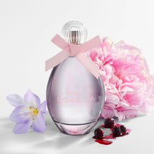 A perfume bottle labeled 'born LOVELY' with a pink ribbon bow, styled with a pink peony flower, a purple iris, and red cherries on a clean white background.