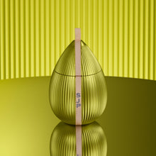 A metallic gold egg-shaped container with vertical ridges and a beige tape measure standing upright inside it, set on a reflective surface against a yellow-green ribbed background.